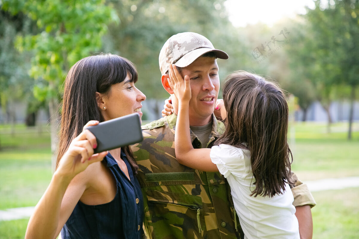 公园快乐的军人享受与妻子和小女儿在一起的时光 在城市公园用手机自拍中镜头家庭团聚或回家的概念白种人制服手机