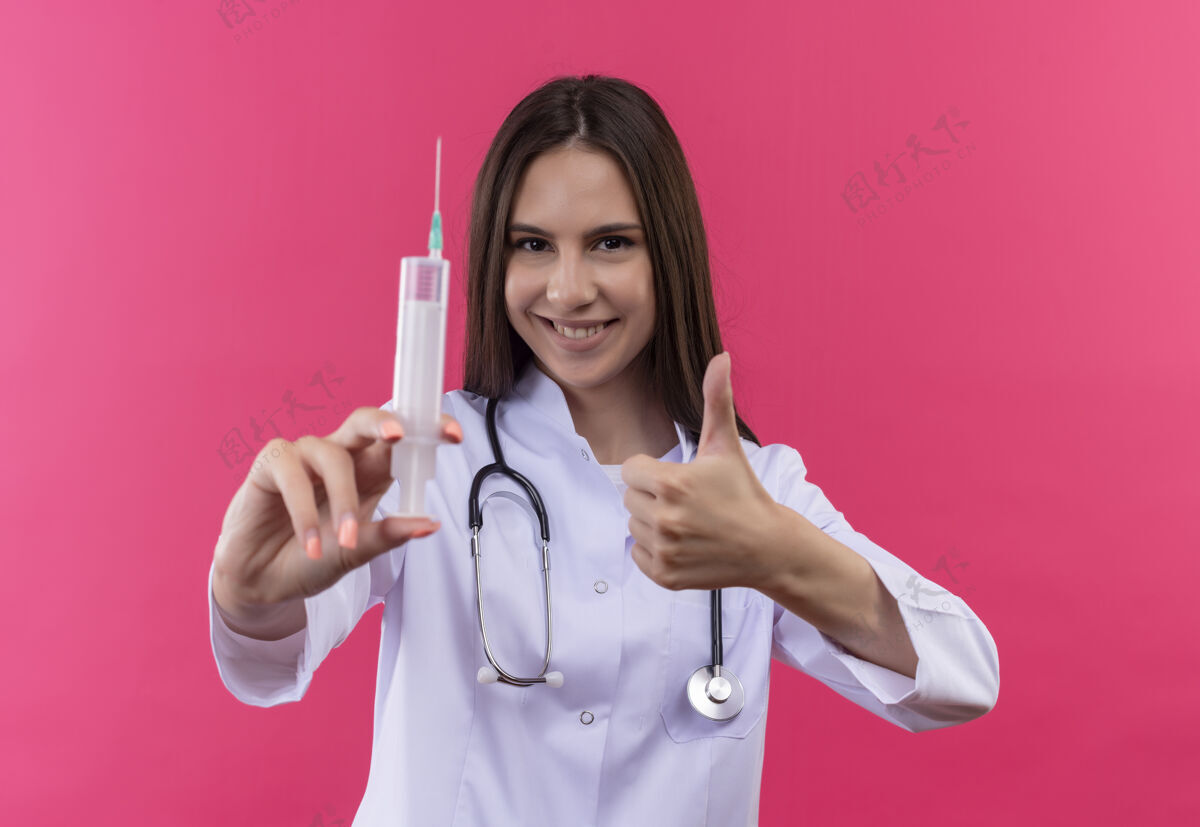 向上微笑的年轻医生女孩穿着听诊器医用长袍拿着注射器 她的大拇指在孤立的粉红色背景上向上女孩医生年轻