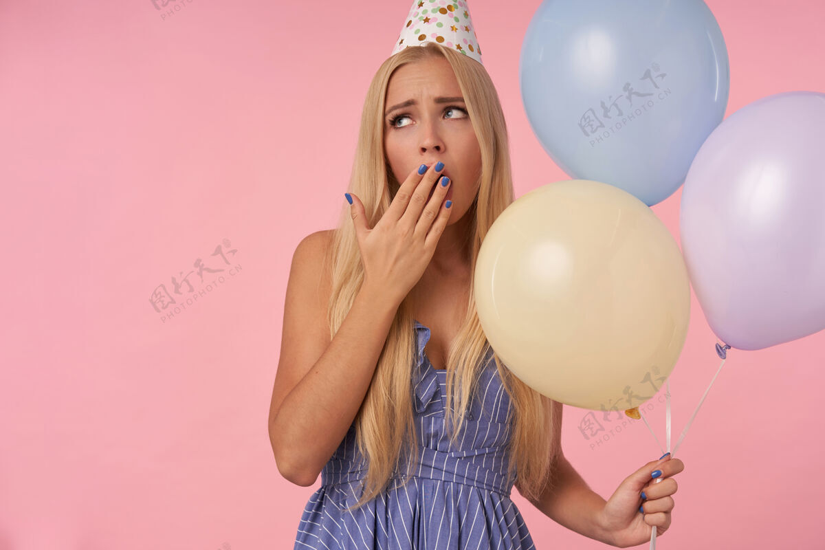 肖像无聊的年轻女性 一头金色长发 穿着节日服装 戴着圆锥形帽子 手里拿着一堆氦气球 在粉色背景上摆姿势 打呵欠 举起手捂着嘴打哈欠室内发型