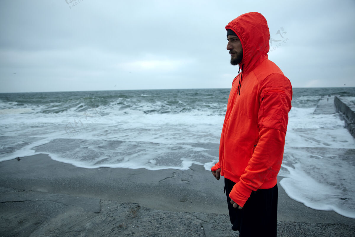 海滨在灰色的清晨 身穿暖橙色连帽衫和黑色运动裤的年轻黑发胡须运动员站在海边 若有所思地向前看户外运动员运动型