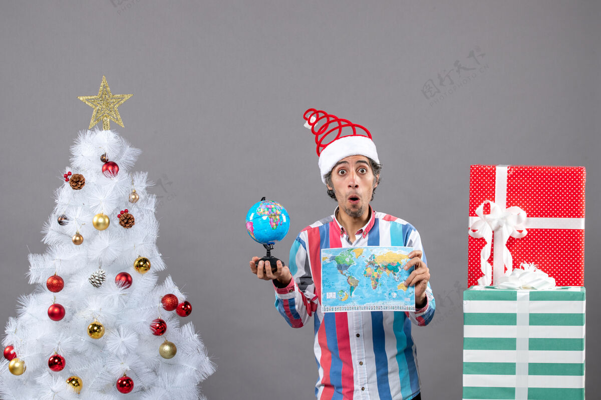 圣诞前视图惊讶的人拿着世界地图和地球仪站在白色圣诞树旁边 星星服装惊讶男人