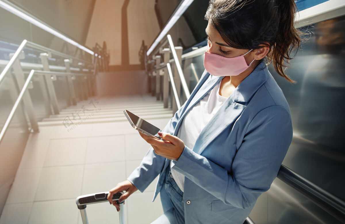 智能手机大流行期间在机场使用智能手机的戴着医用面罩和行李的高角度女性Covid19冠状病毒楼梯