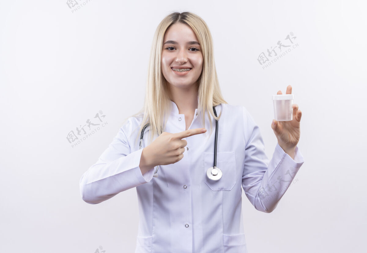 女孩微笑的医生年轻的金发女孩戴着听诊器和医用长袍戴着牙套指着空罐 手上拿着孤立的白色背景支架长袍持有