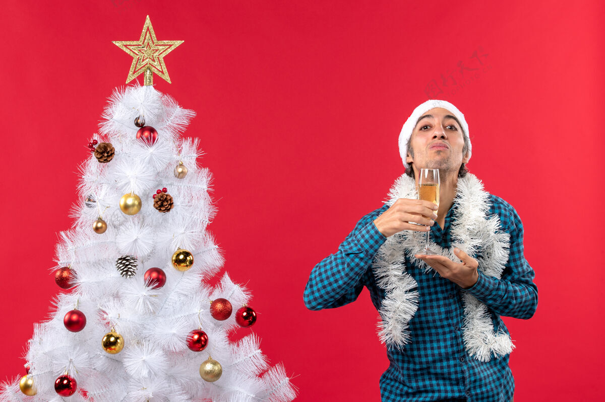 装饰圣诞节心情与情绪激动的年轻人圣诞老人帽子在一个蓝色的剥离衬衫举行一杯葡萄酒在他的下巴附近圣诞树年轻人年搞笑