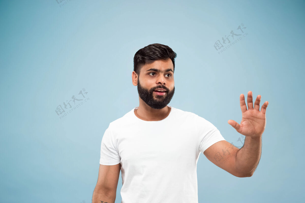 工作蓝色背景上穿着白衬衫的印度教年轻人的半身特写肖像人类情感 面部表情 广告概念负面空间显示空白栏 指向 问候公司高管顺序