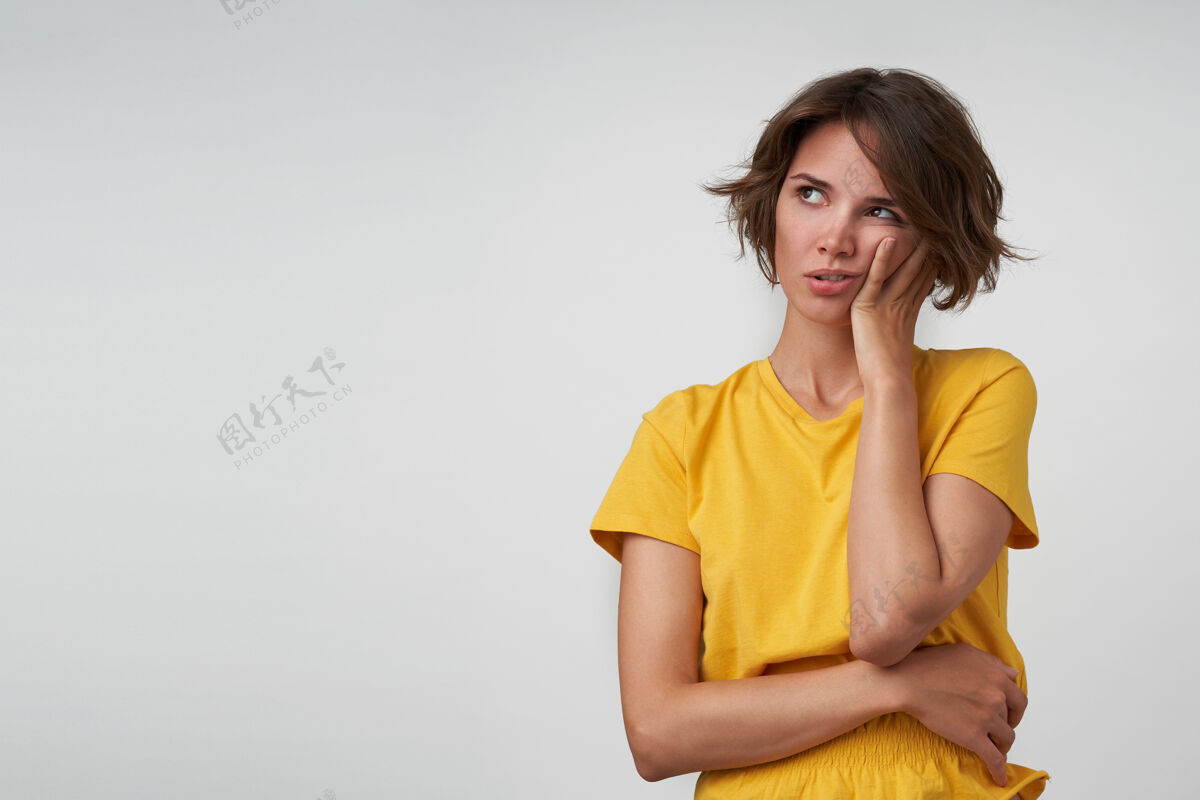 姿势迷惑不解的年轻漂亮女性 棕色短发把脸颊放在抬起的手上 一边看一边 一边摆姿势 一边穿着黄色t恤穿着上衣心情