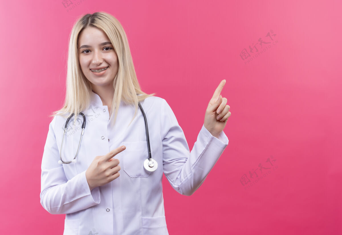 金发微笑的医生年轻的金发女孩穿着医用长袍戴着听诊器和牙套指向一边 双手放在孤立的粉红色背景上牙科穿着听诊器