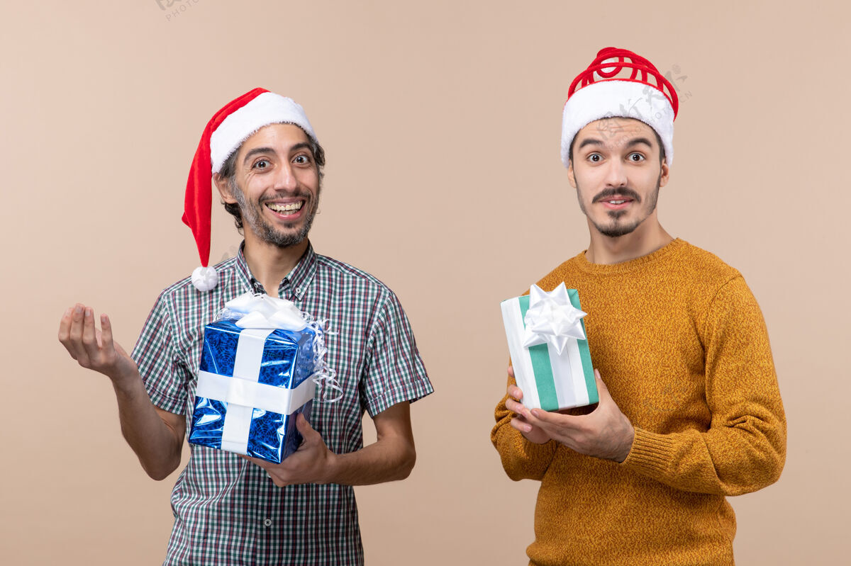 两个微笑的男人正面图两个微笑的家伙戴着圣诞帽 拿着圣诞礼物 背景是米色的礼物风景帽子
