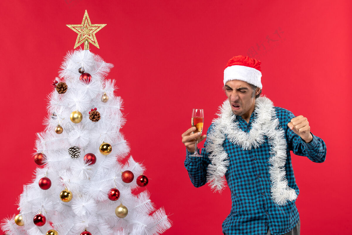 新圣诞心情与骄傲的年轻人圣诞老人帽子在一个蓝色条纹衬衫举起一杯葡萄酒显示他的幸福圣诞树附近帽子心情新年前夜