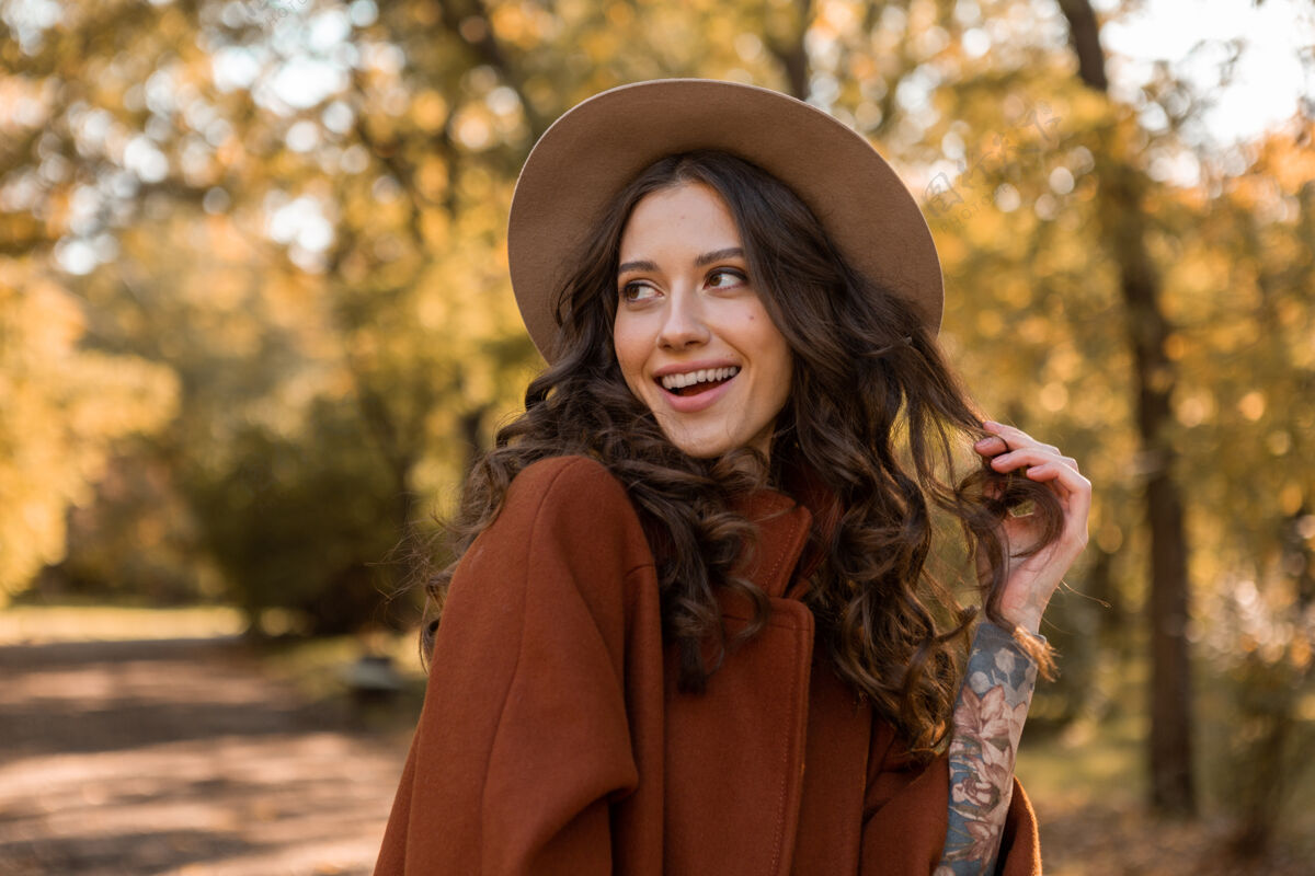 欢快画中有魅力的时尚微笑女子留着长长的卷发漫步在公园里穿着温暖的棕色外套秋日时尚 街头风格戴着帽子苗条休闲漂亮