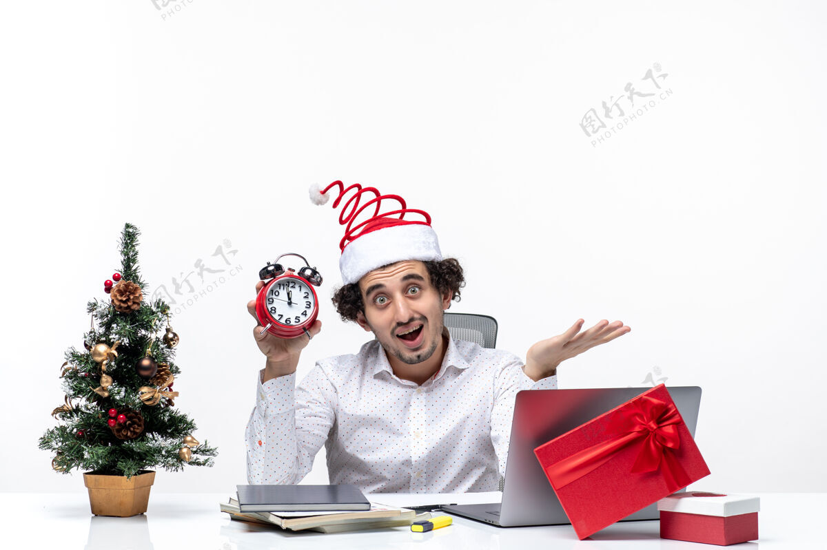 圣诞老人戴着圣诞老人帽 拿着时钟 在白色背景上指着办公室里的东西的年轻快乐的商人什么的圣诞节微笑