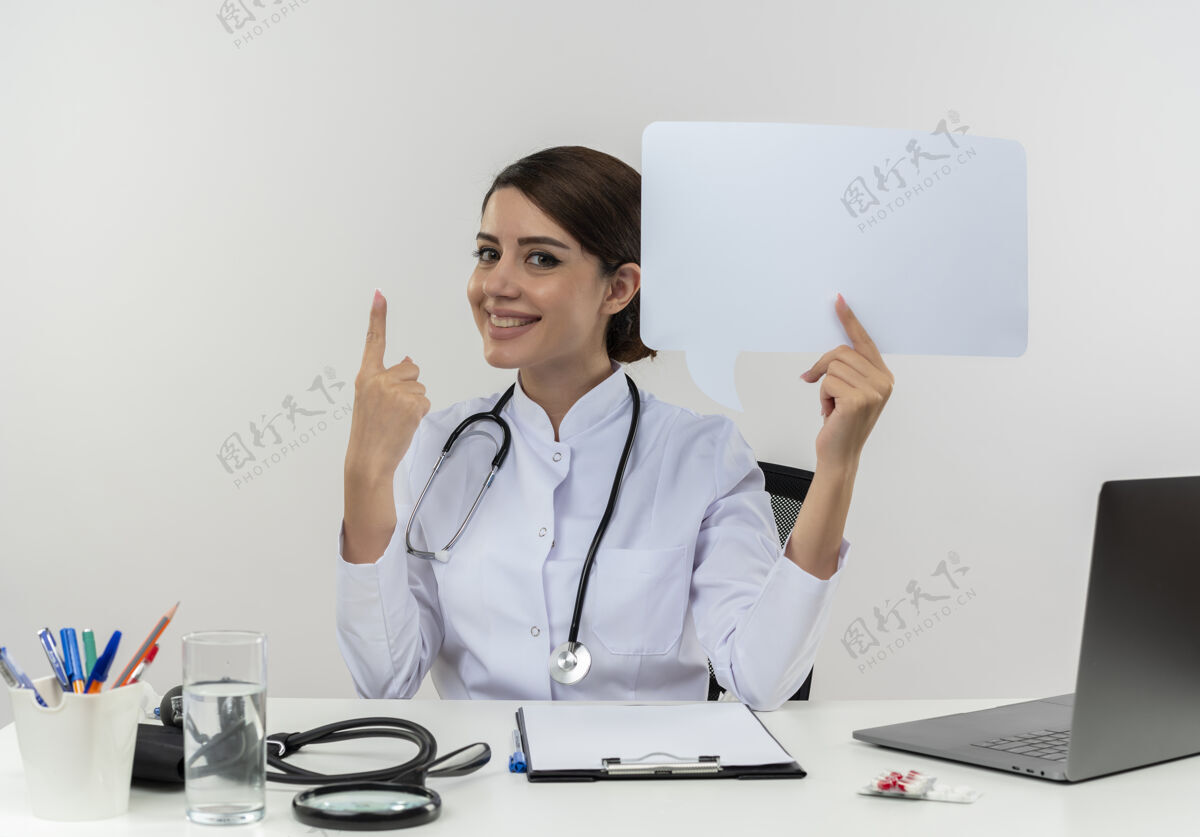 坐着面带微笑的年轻女医生穿着医用长袍 手持听诊器坐在办公桌旁 拿着医疗工具 拿着聊天泡泡指向上的复印空间电脑听诊器拿着