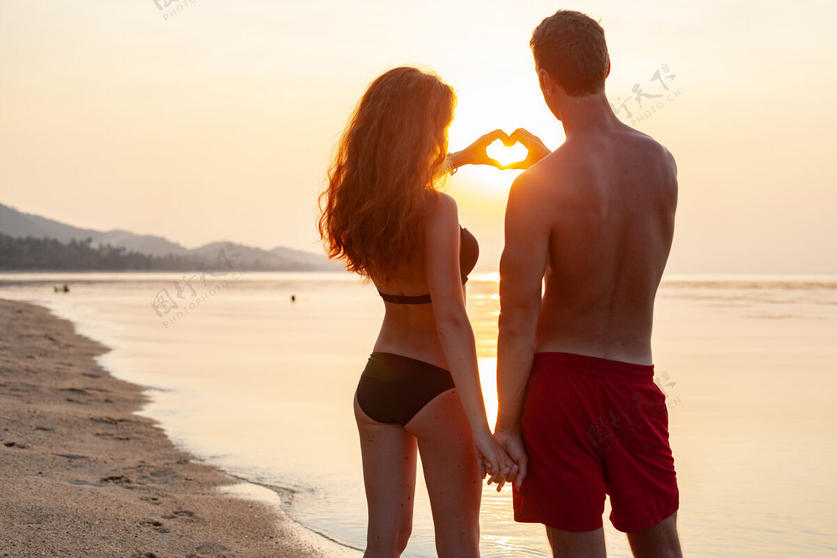 夏天年轻性感浪漫的情侣在夏天的沙滩上快乐地在一起 穿着泳衣在日落时露出心形标志户外海滩热带