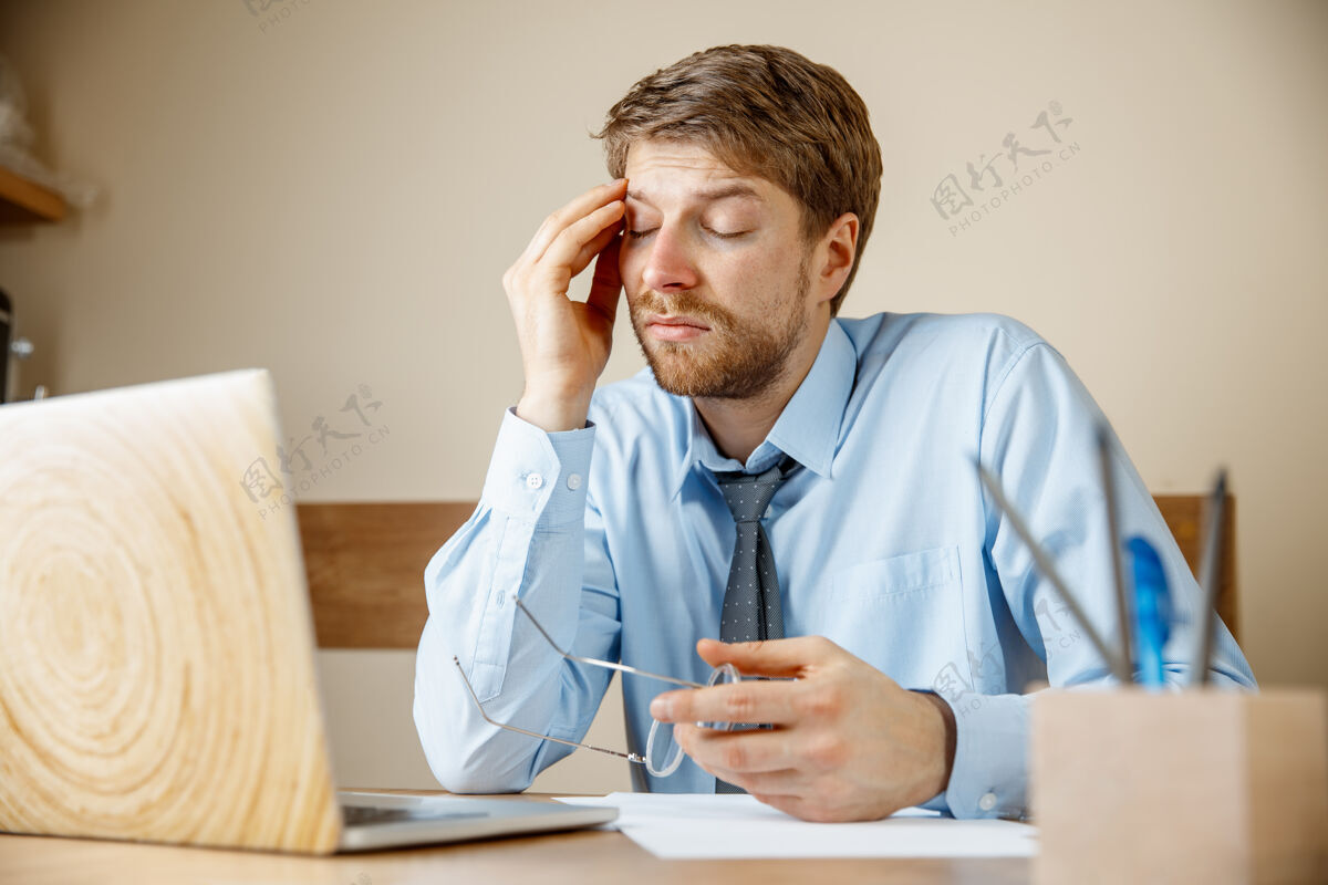 专业感觉不舒服和疲倦沮丧悲伤不快乐的生病的年轻人坐在办公室工作时按摩他的头季节性流感 大流行性流感 疾病预防概念压力笔记本电脑忙碌
