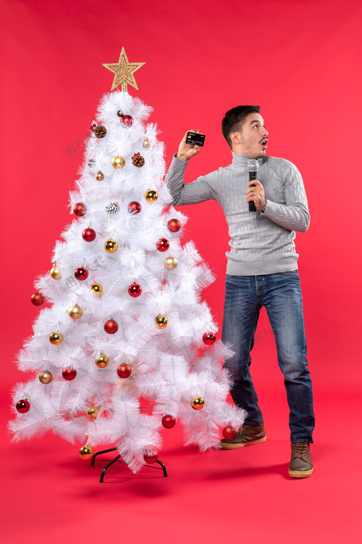 拿着圣诞节的心情与自信的家伙站在装饰圣诞树附近 拿着麦克风拍照 看东西站着看着左边