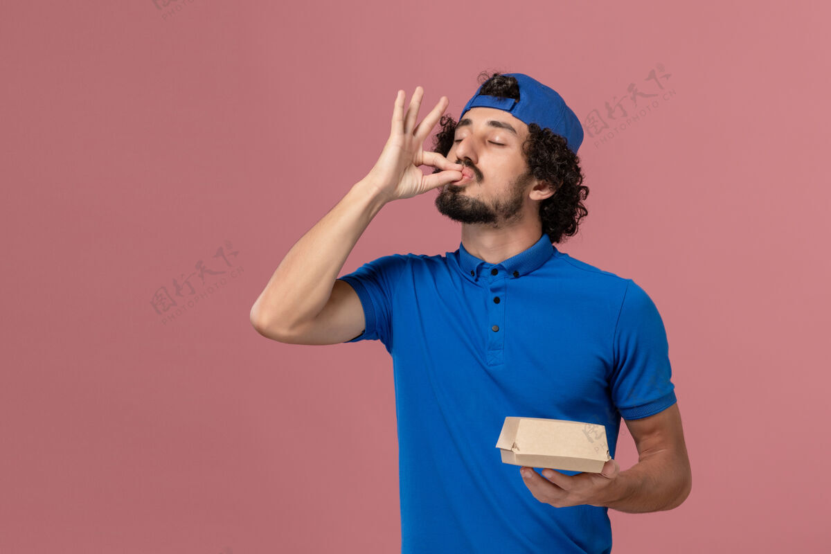 人正面图身穿蓝色制服和披风的男性快递员拿着粉色墙上的小快递食品包食物视图粉色