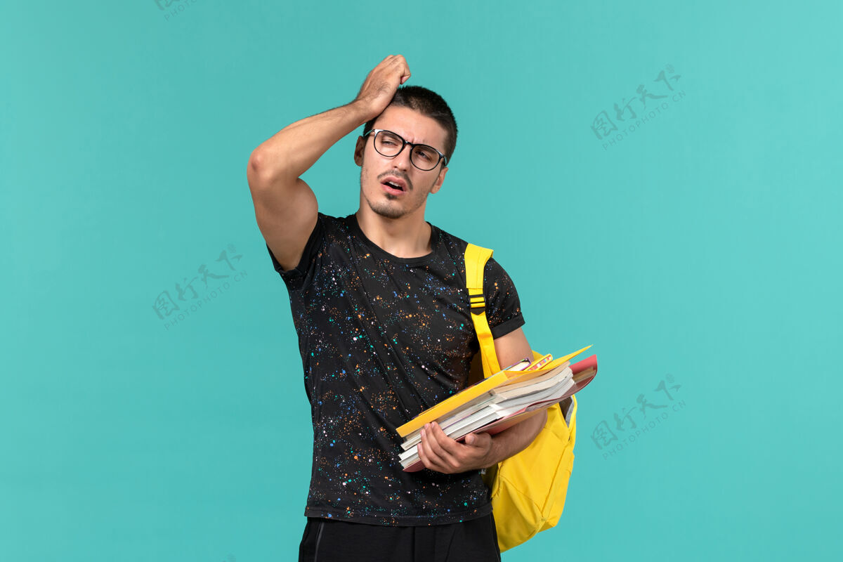 书身穿深色t恤黄色背包的男学生正面图 蓝色墙上有文件和书籍持有学校男性