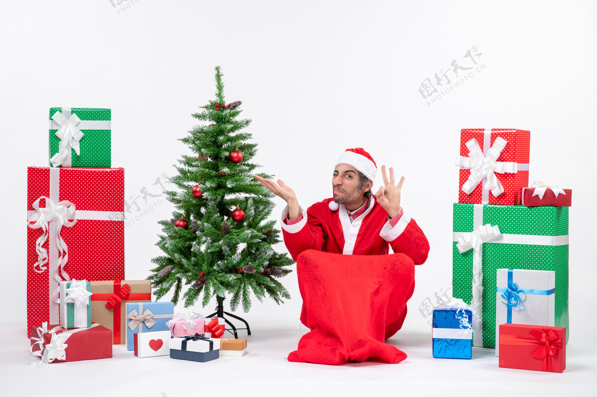 眼镜一脸酸酸的年轻人打扮成圣诞老人 拿着礼物和装饰好的圣诞树坐在地上做着戴眼镜的手势 指着白色背景上的东西圣诞手势指向