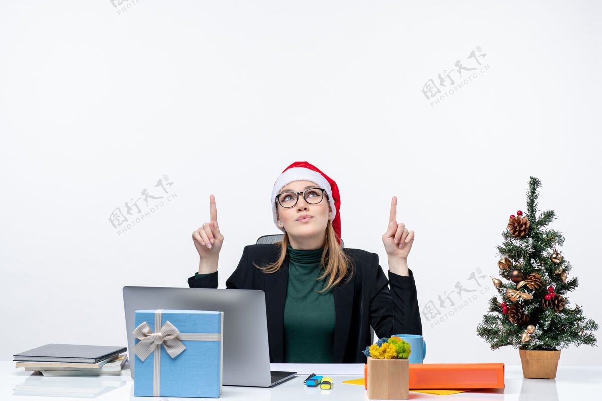 礼物一位积极的商务女性 戴着圣诞老人帽 坐在一张桌子旁 桌子上有一棵圣诞树 上面有一份礼物 背景是白色的职业电脑办公室