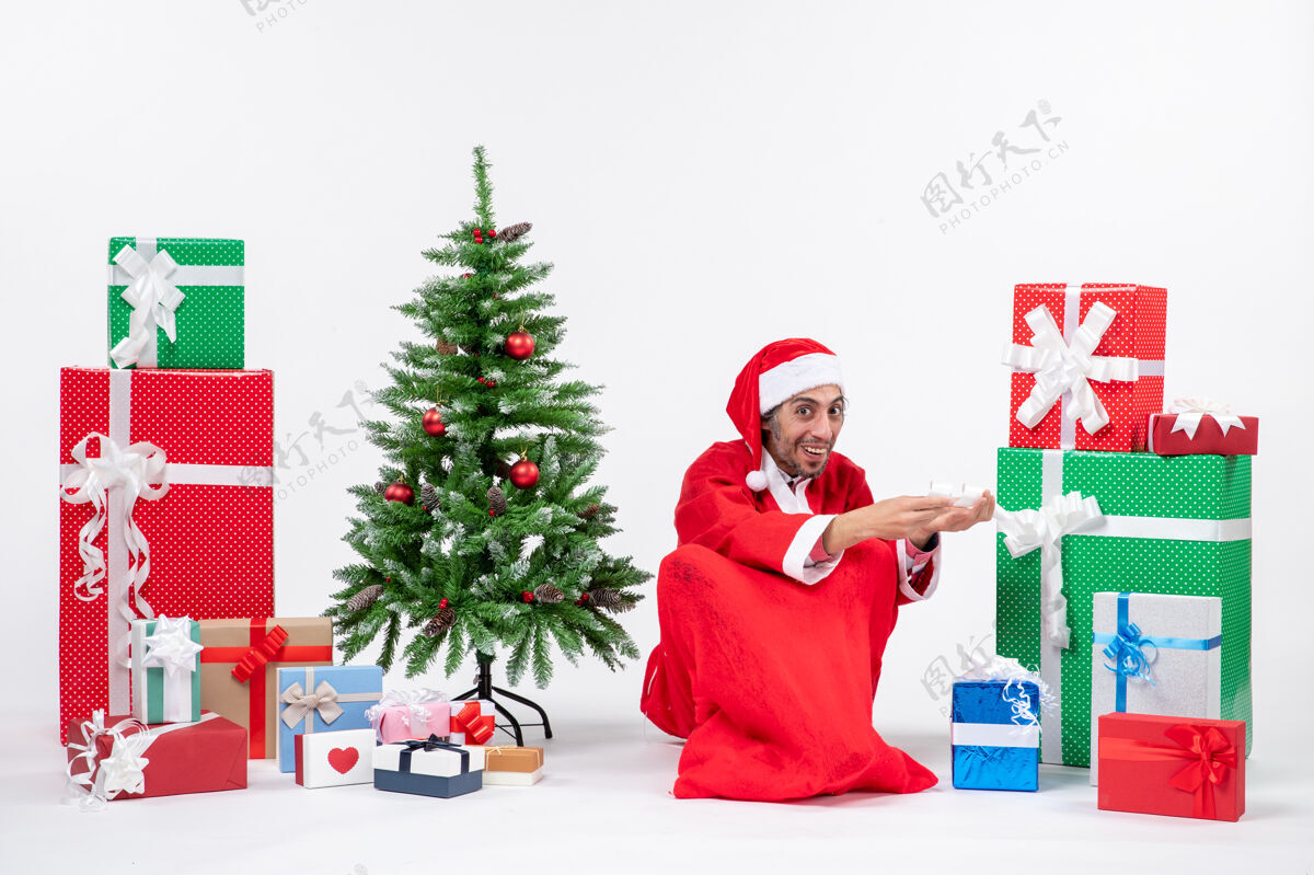 微笑微笑着惊讶的年轻人打扮成圣诞老人和礼物装饰圣诞树坐在地上的白色背景圣诞老人人礼物