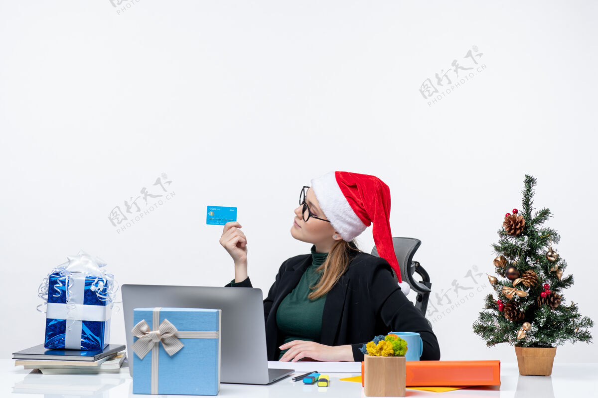 有吸引力自信 积极 忙碌 有魅力的女人戴着圣诞老人的帽子 戴着眼镜坐在桌子旁 手里拿着银行卡在办公室里电脑圣诞节眼镜