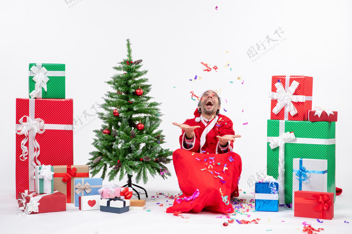 圣诞树欢乐的圣诞老人坐在地上 在礼物和白色背景装饰圣诞树旁玩圣诞装饰品 带来节日的气氛圣诞坐礼物