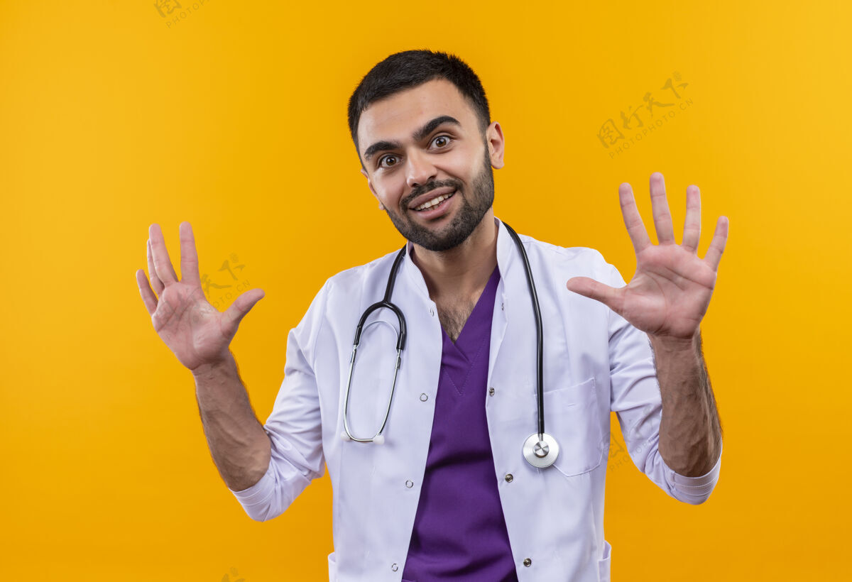 穿着微笑的年轻男医生穿着听诊器医用长袍 在孤立的黄色背景上举手听诊器长袍提高