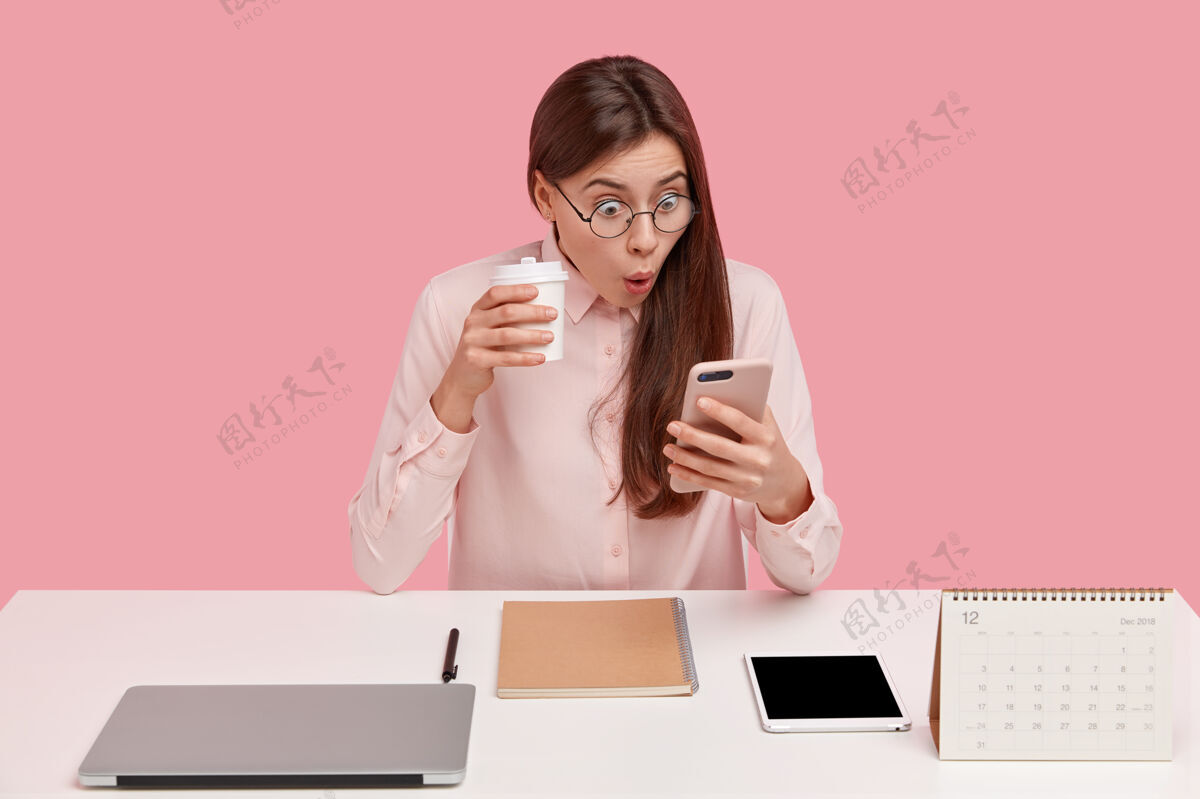 惊讶一张漂亮的年轻女子的照片 在手机上阅读令人震惊的新闻 在社交网络上观看视频 用一次性杯子喝咖啡学生浏览咖啡