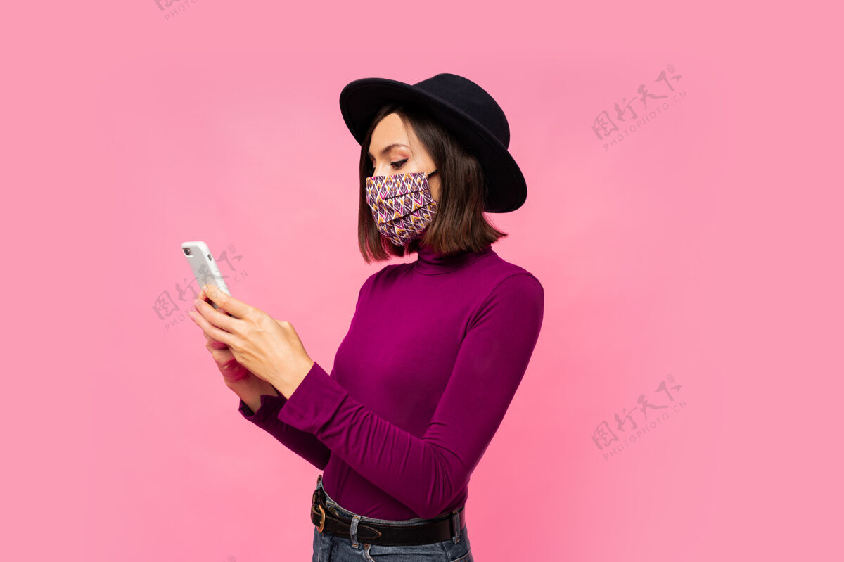 保护戴着防护面具和黑帽子的时髦女人用手机 站着面罩拿着女性