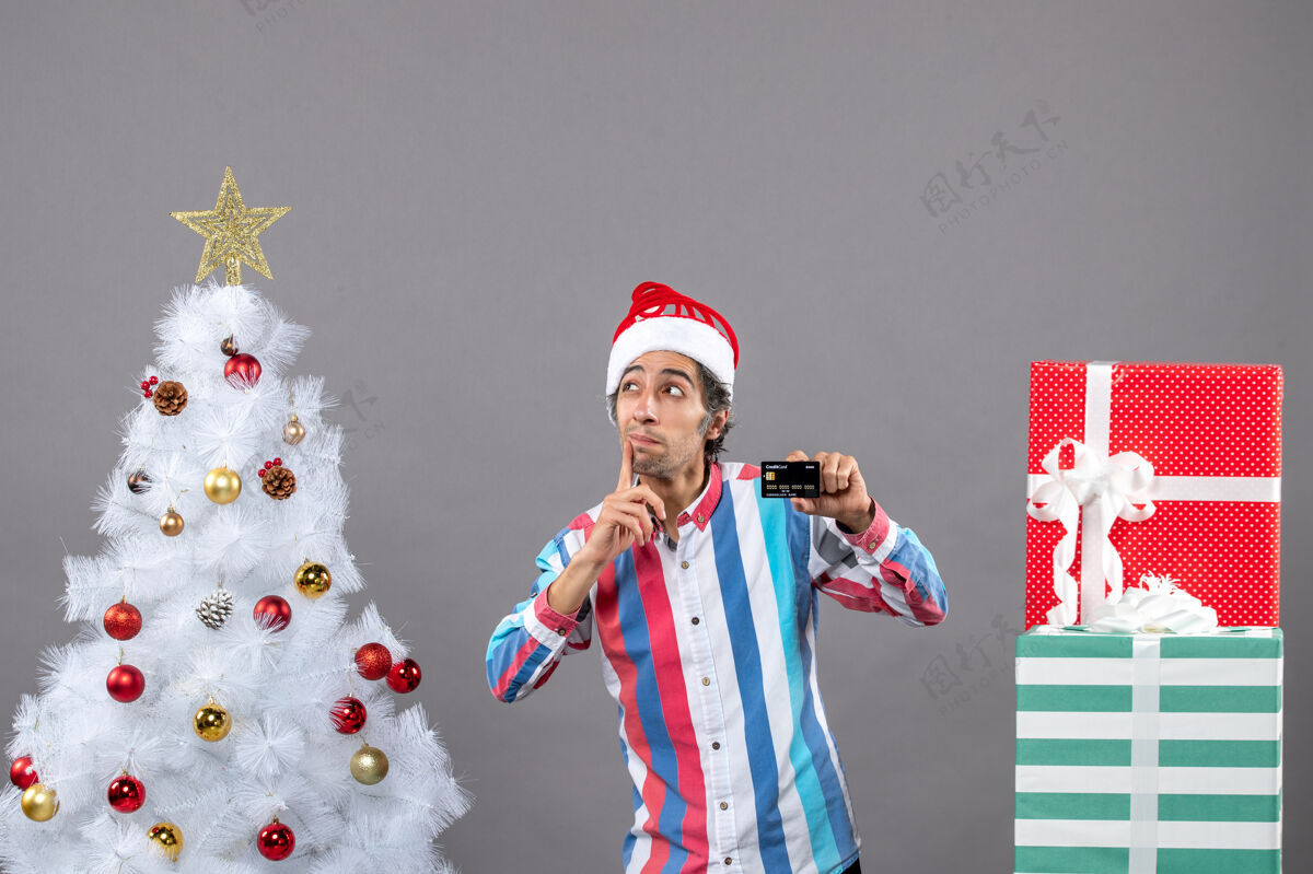 站着正面图好奇的男人拿着信用卡把手放在脸颊上站在不同的礼物旁边好奇的人肖像男性