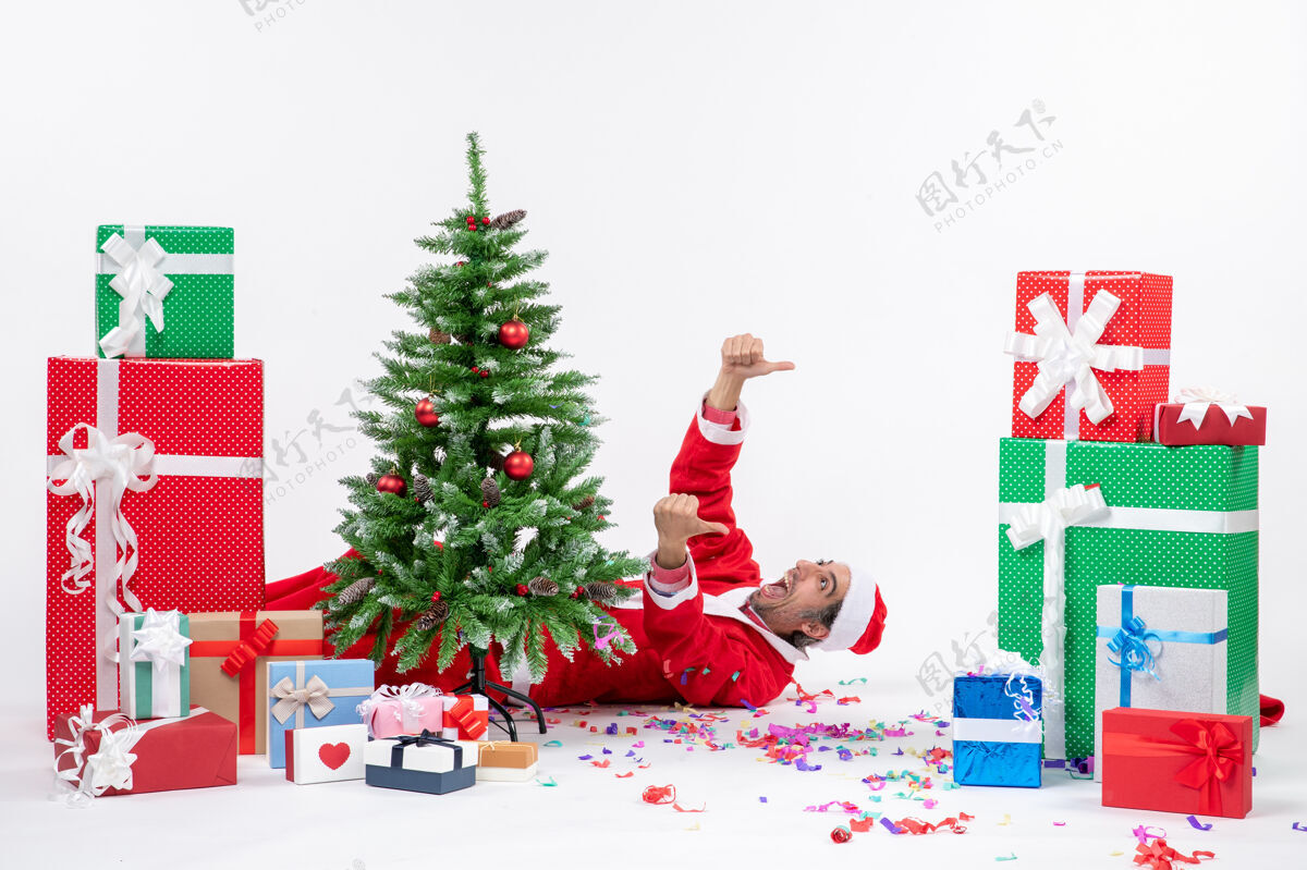 礼物节日气氛与年轻积极的圣诞老人躺在圣诞树后靠近礼物白色背景圣诞圣诞老人喜庆