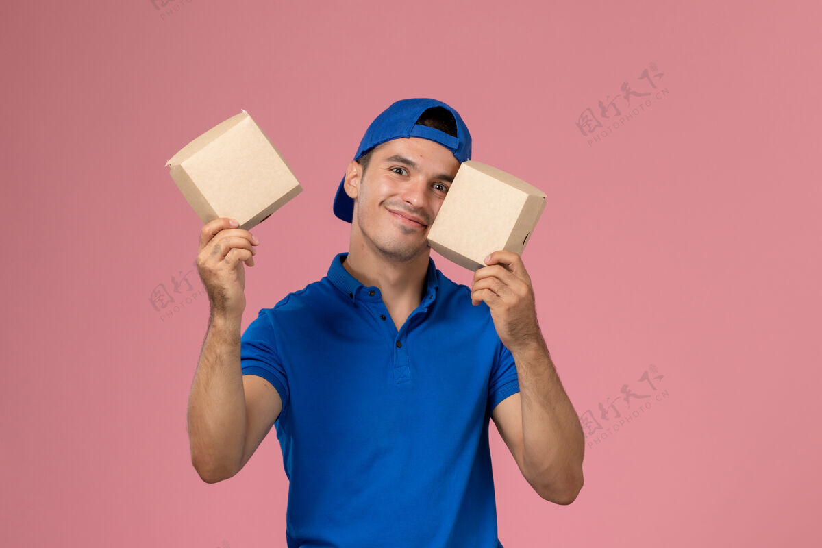服务正面图：身穿蓝色制服披肩的年轻男性快递员手拿着小食品包 站在浅粉色的墙上手持男性包裹