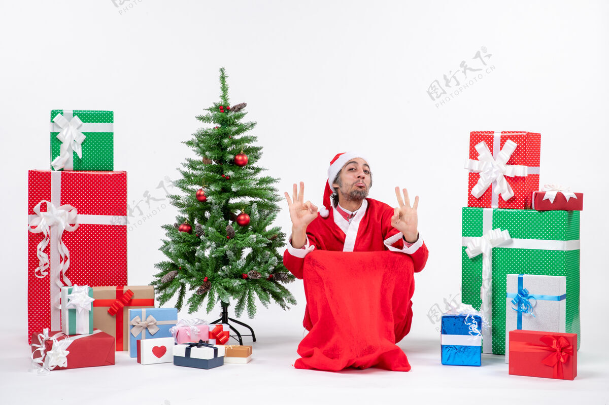 礼物情绪激动的年轻人装扮成圣诞老人 带着礼物和装饰圣诞树 在白色背景上做眼镜手势圣诞老人情绪化圣诞老人