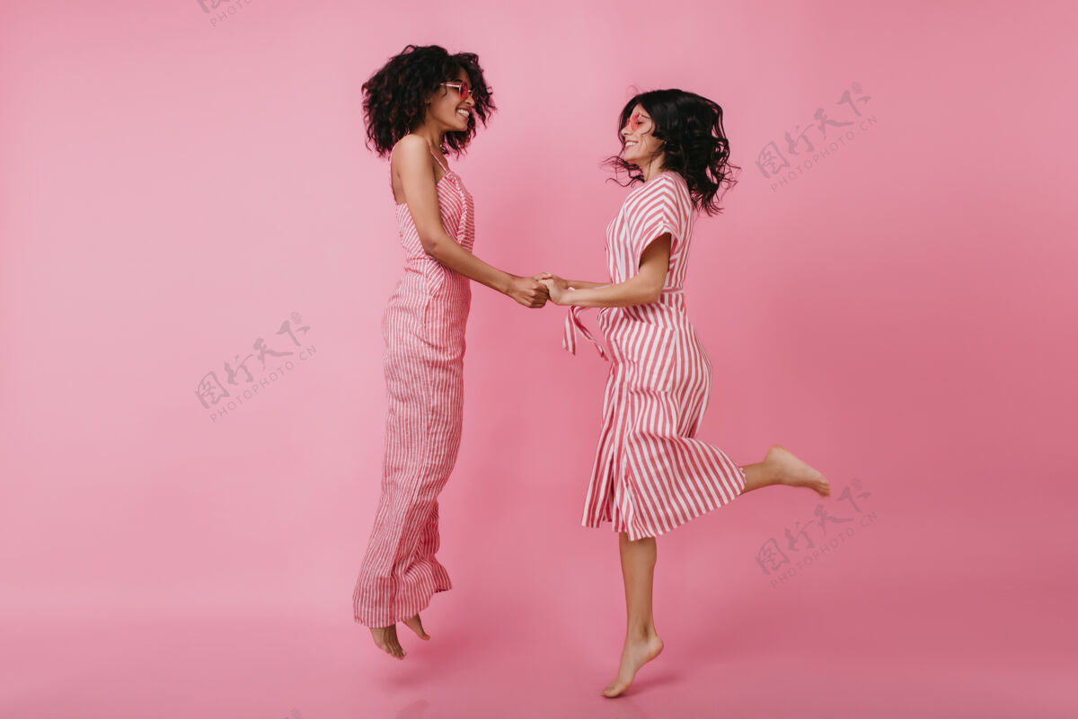 模特卷发的非洲女孩跳起来和朋友牵手穿着粉色连衣裙的无忧无虑的女模特令人毛骨悚然休闲室内舞蹈