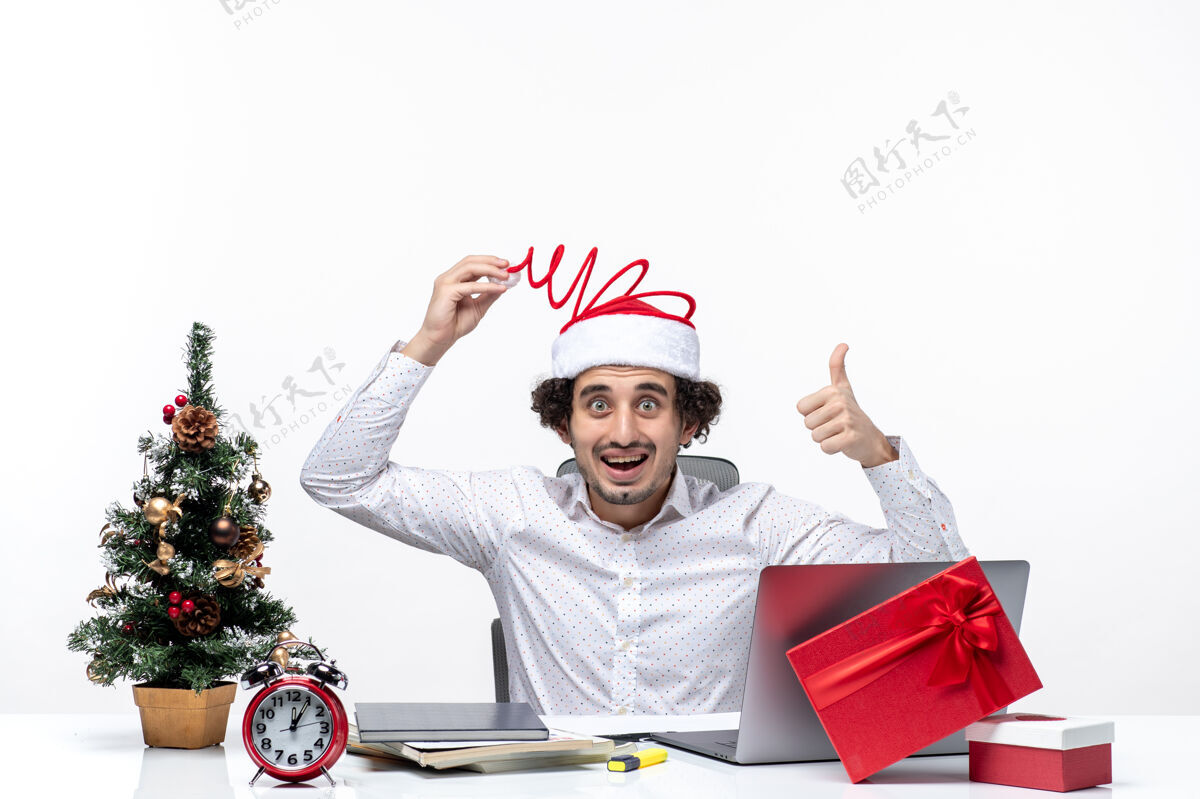 圣诞老人新的一年的心情与惊讶微笑的年轻商人触摸他有趣的圣诞老人帽子指着上面在白色背景的办公室年轻的商人商人办公室