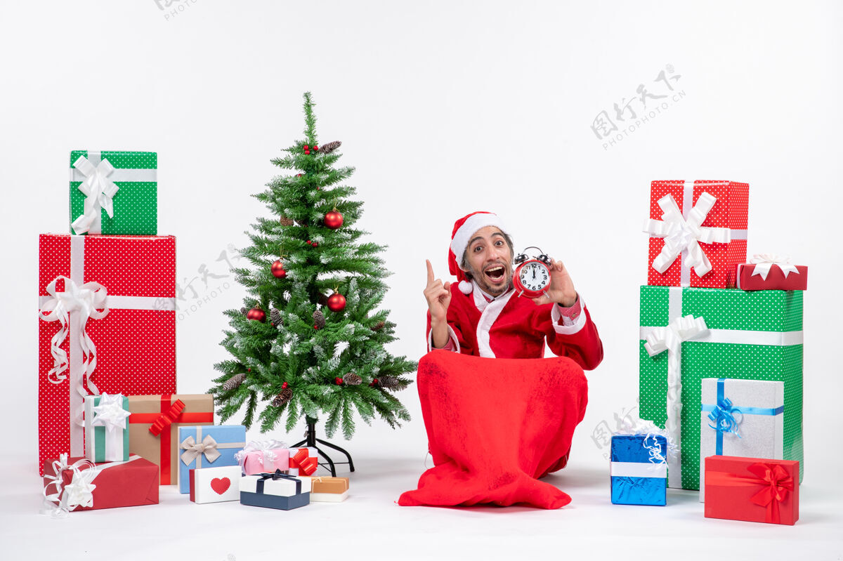 圣诞节好奇的年轻人坐在地上 拿着钟在礼物和圣诞树旁庆祝新年或圣诞节地男人礼物