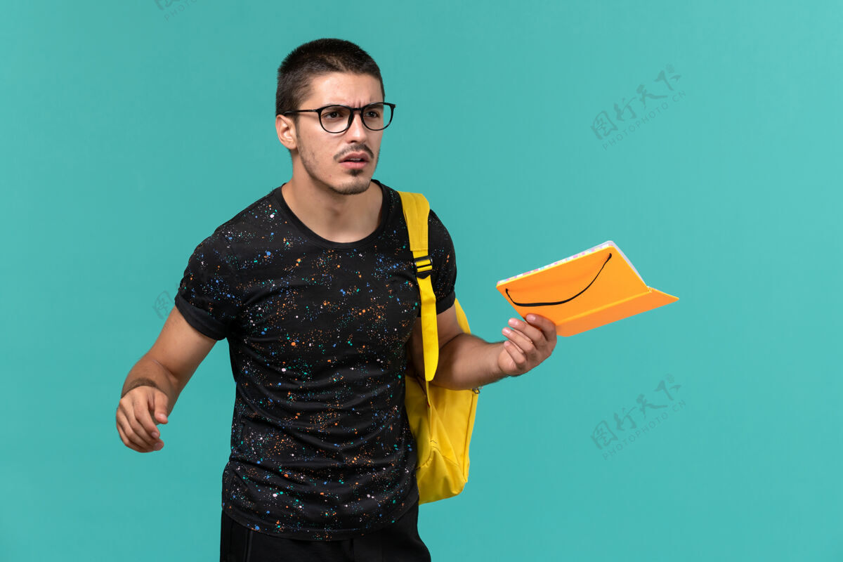 模特身穿深色t恤的男学生正面图黄色背包 蓝色墙上拿着抄写本肖像大学持有