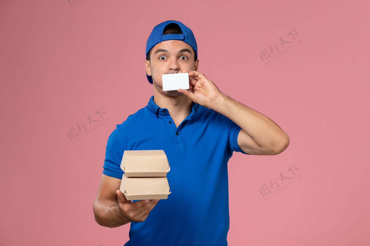 快递正面图：身穿蓝色制服披肩的年轻男性快递员拿着小包裹 浅粉色墙上有卡片人食物成人