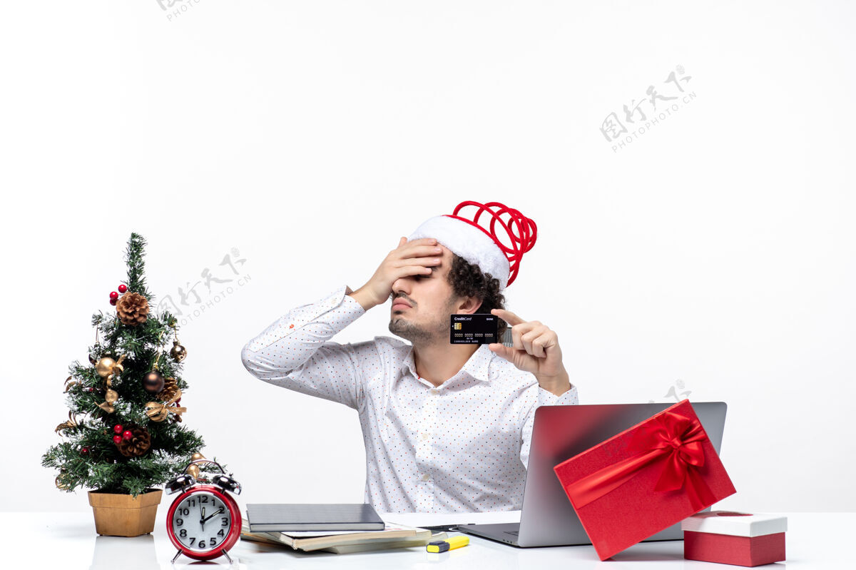 办公室圣诞节心情与年轻的胡子疲惫的商人与圣诞老人帽子持有银行卡 并指出在办公室以上指着微笑年轻人