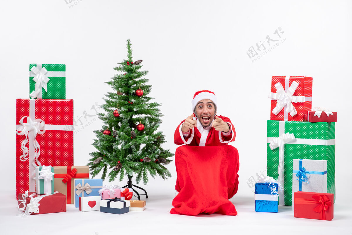 年轻人情绪激动的年轻人打扮成圣诞老人与礼物和装饰圣诞树指向白色背景上的东西礼物打扮圣诞老人