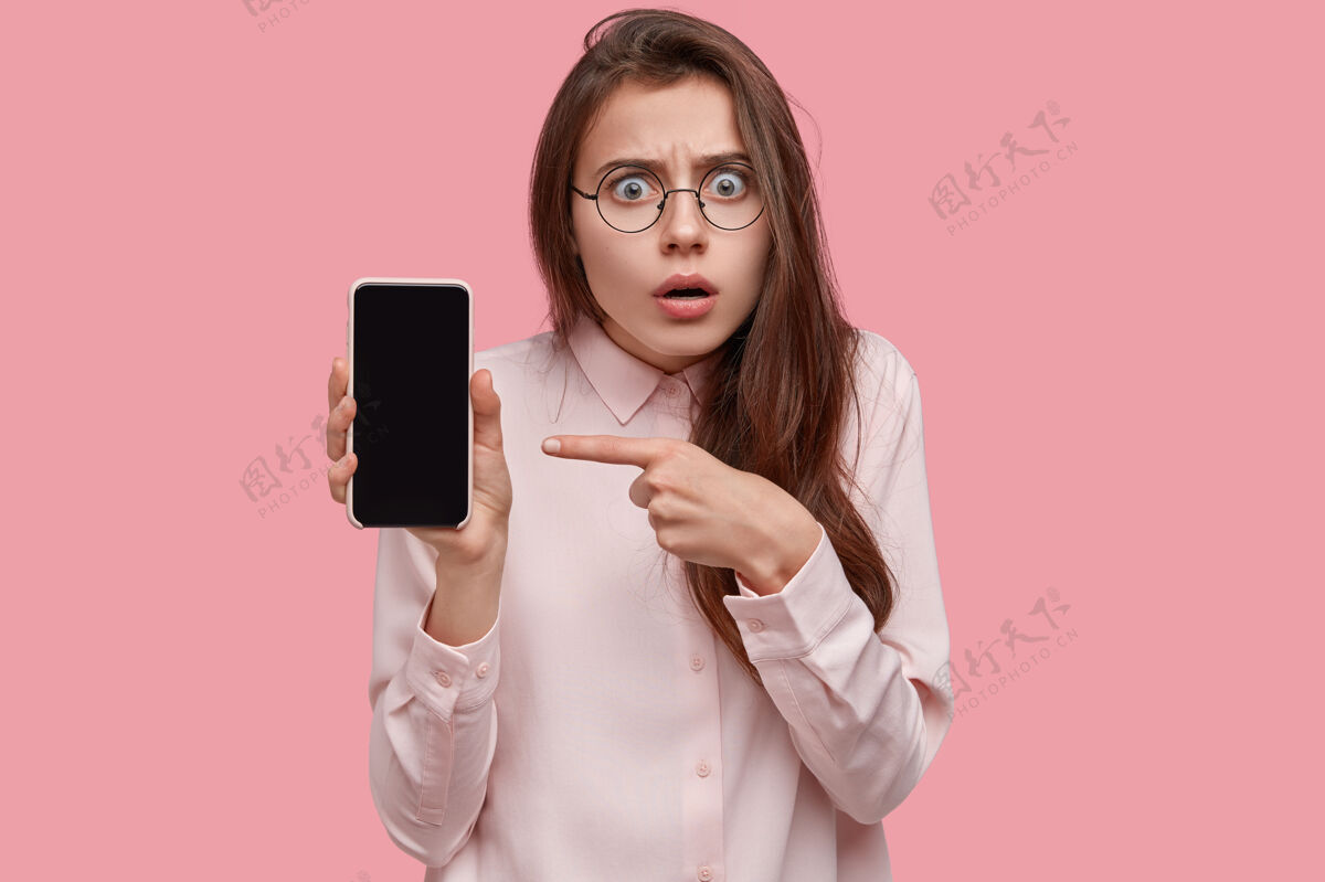 女性惊呆了的黑发女人拿着黑屏智能手机 指着手机 被巨大的折扣惊呆了 试着理解一些东西震惊消息屏幕