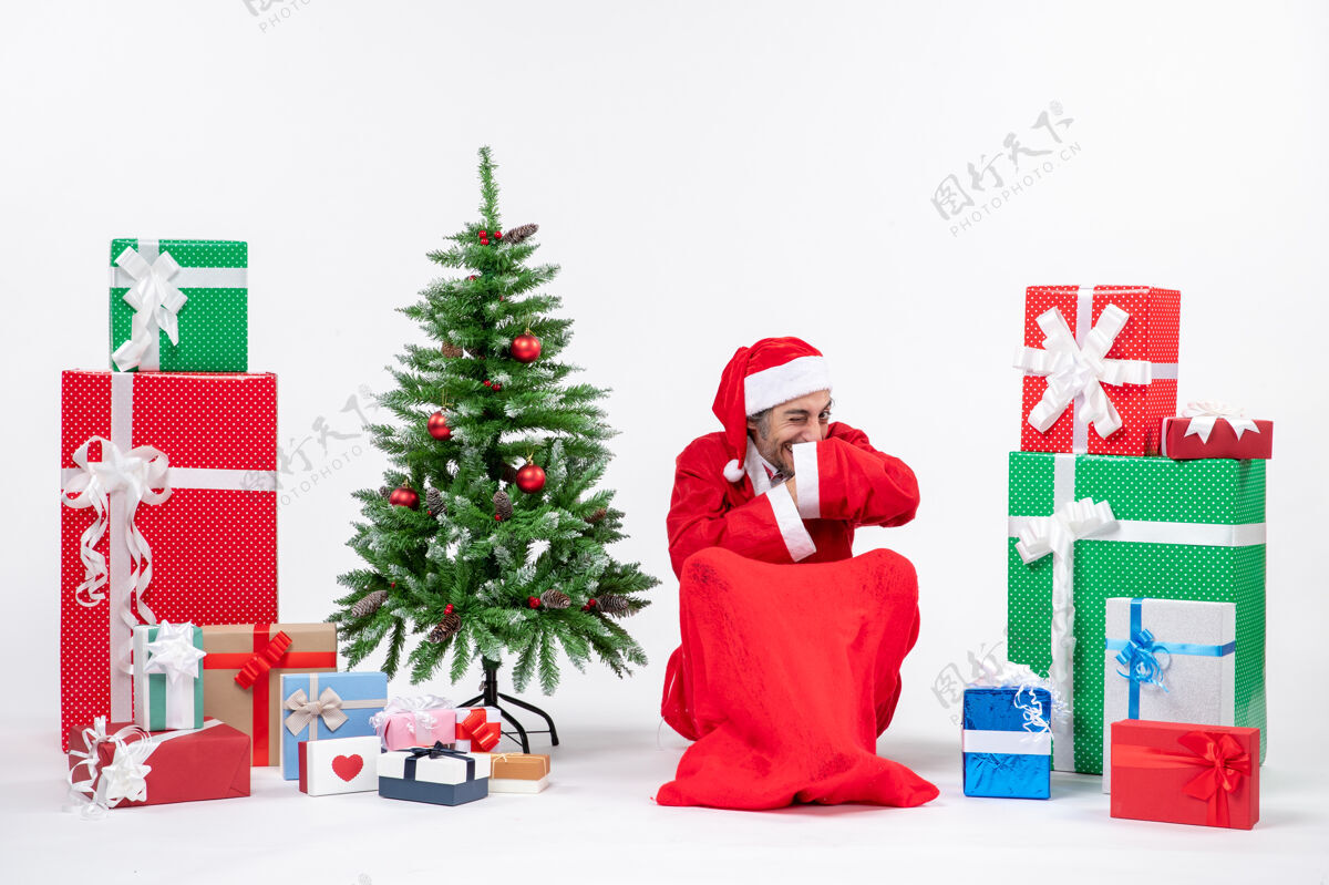 坐着快乐的年轻人带着滑稽的表情装扮成圣诞老人 拿着礼物和装饰好的圣诞树坐在白色背景的地上圣诞装饰打扮