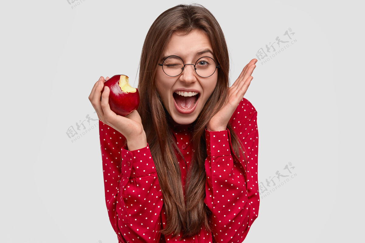 维生素高兴的年轻女子头像眨着眼睛 举手靠近头部 咬着新鲜的苹果 有喜悦的表情 穿着红色圆点衬衫眼镜高兴女性