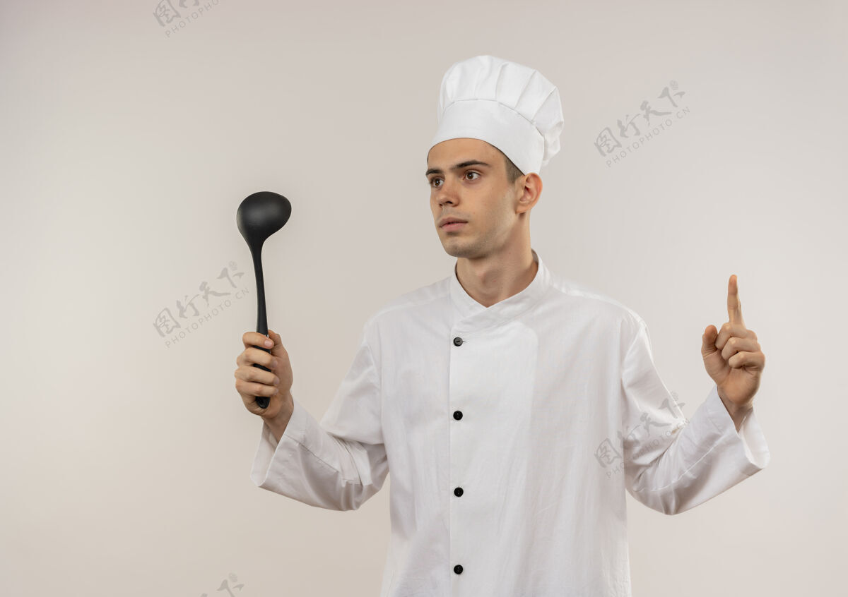 看看着旁边年轻的男厨师穿着厨师制服 拿着勺子指着孤立的白墙上的手指厨师制服侧