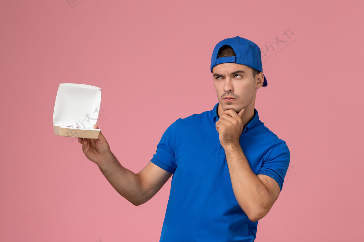 男人正面图身穿蓝色制服斗篷的年轻男性信使手拿着一个小小的空送食品的包裹在粉色的墙上思考粉色服务空