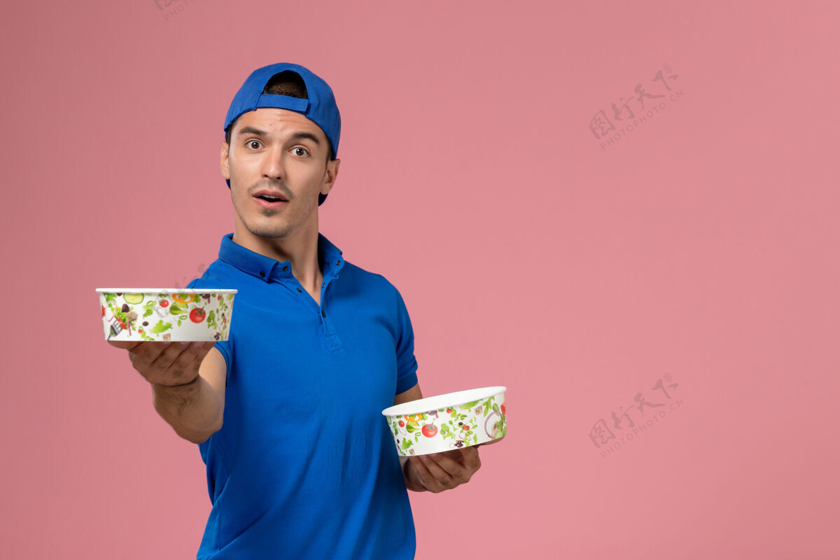 男士正面图：身穿蓝色制服披肩的年轻男性信使手持圆形的投递碗站在浅粉色的墙上专业成人快递员